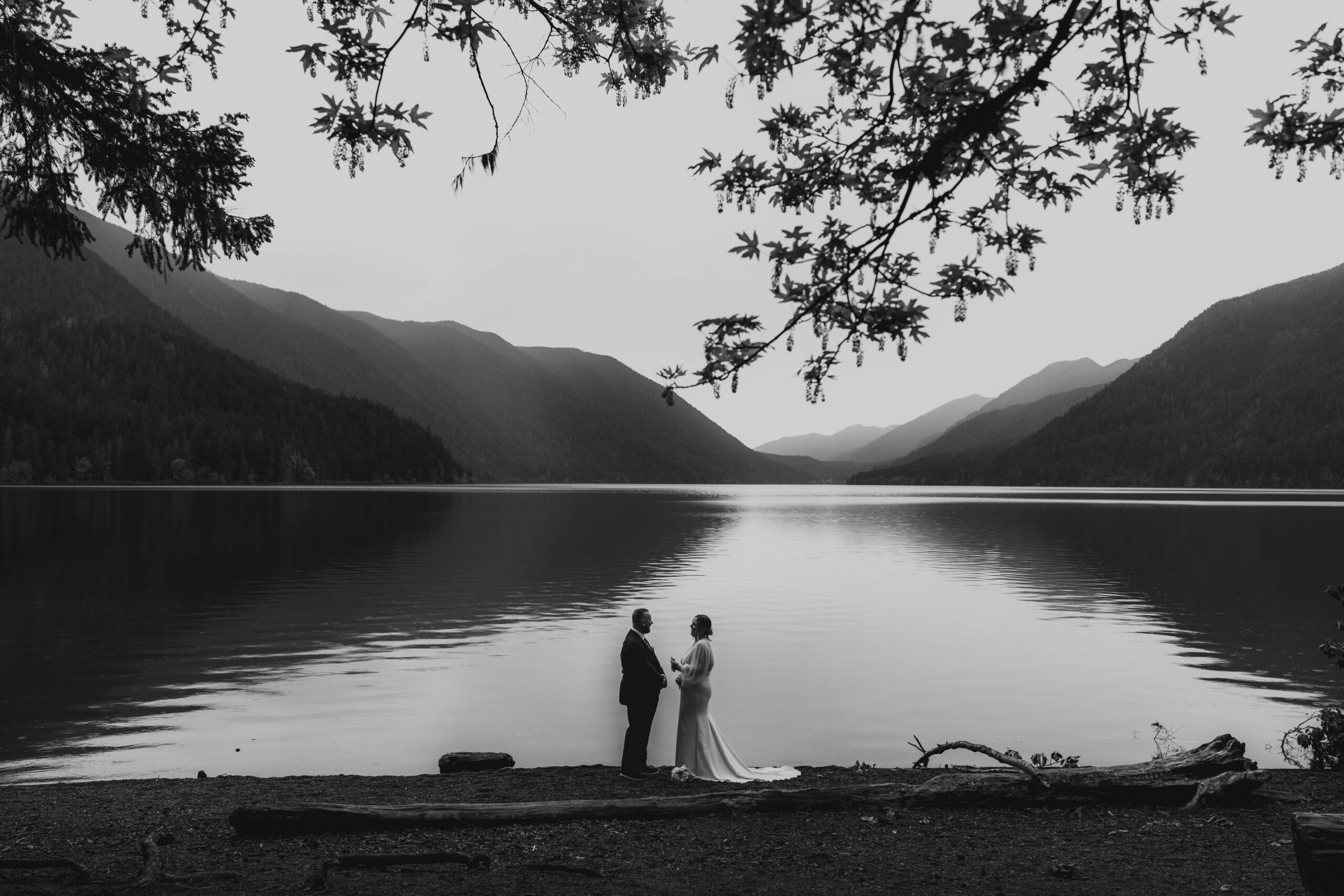 black and white of couple in a distance in wedding attire reading private vows with lake and mountain backdrop
