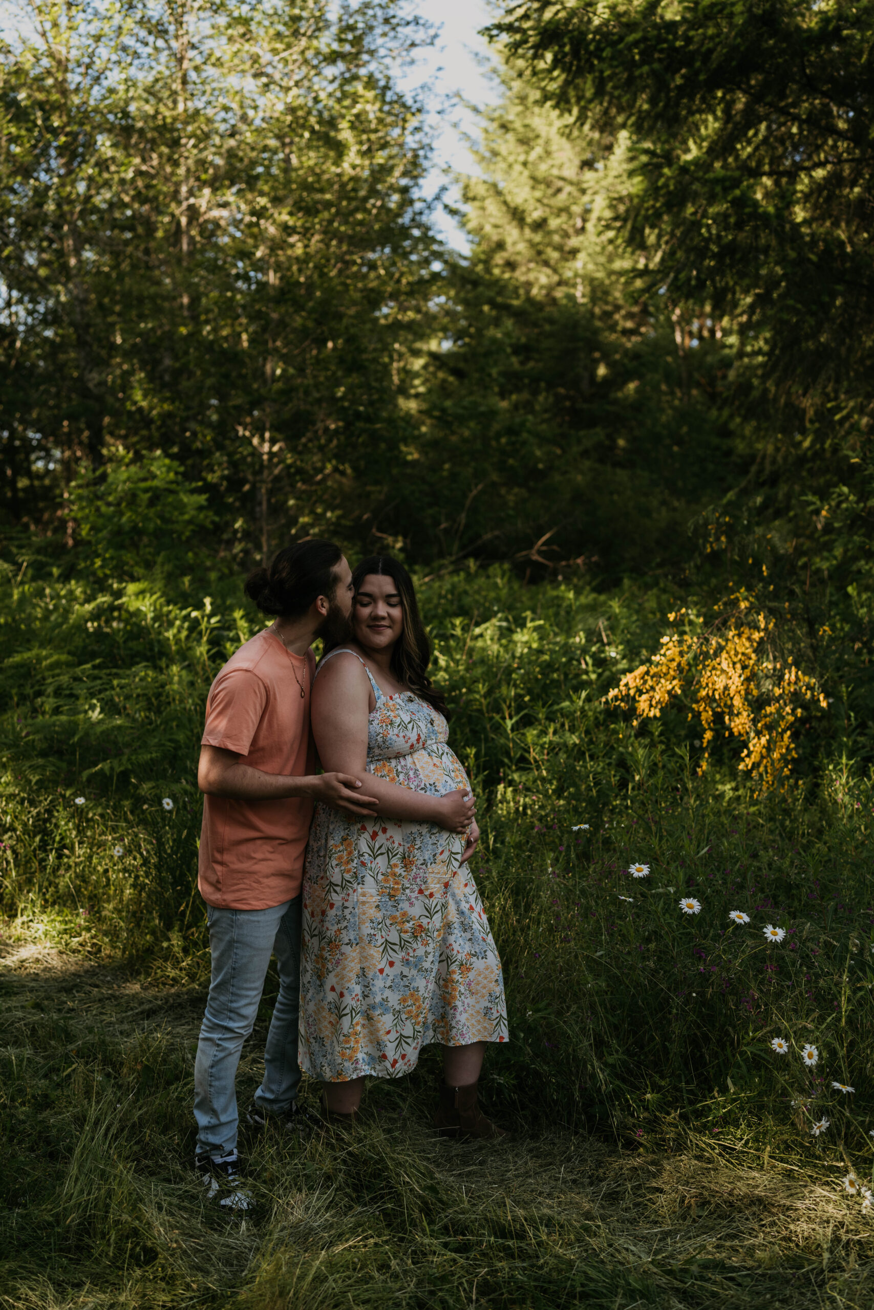couple in forest for maternity session man holding women and women holding belly, man giving her kiss on head