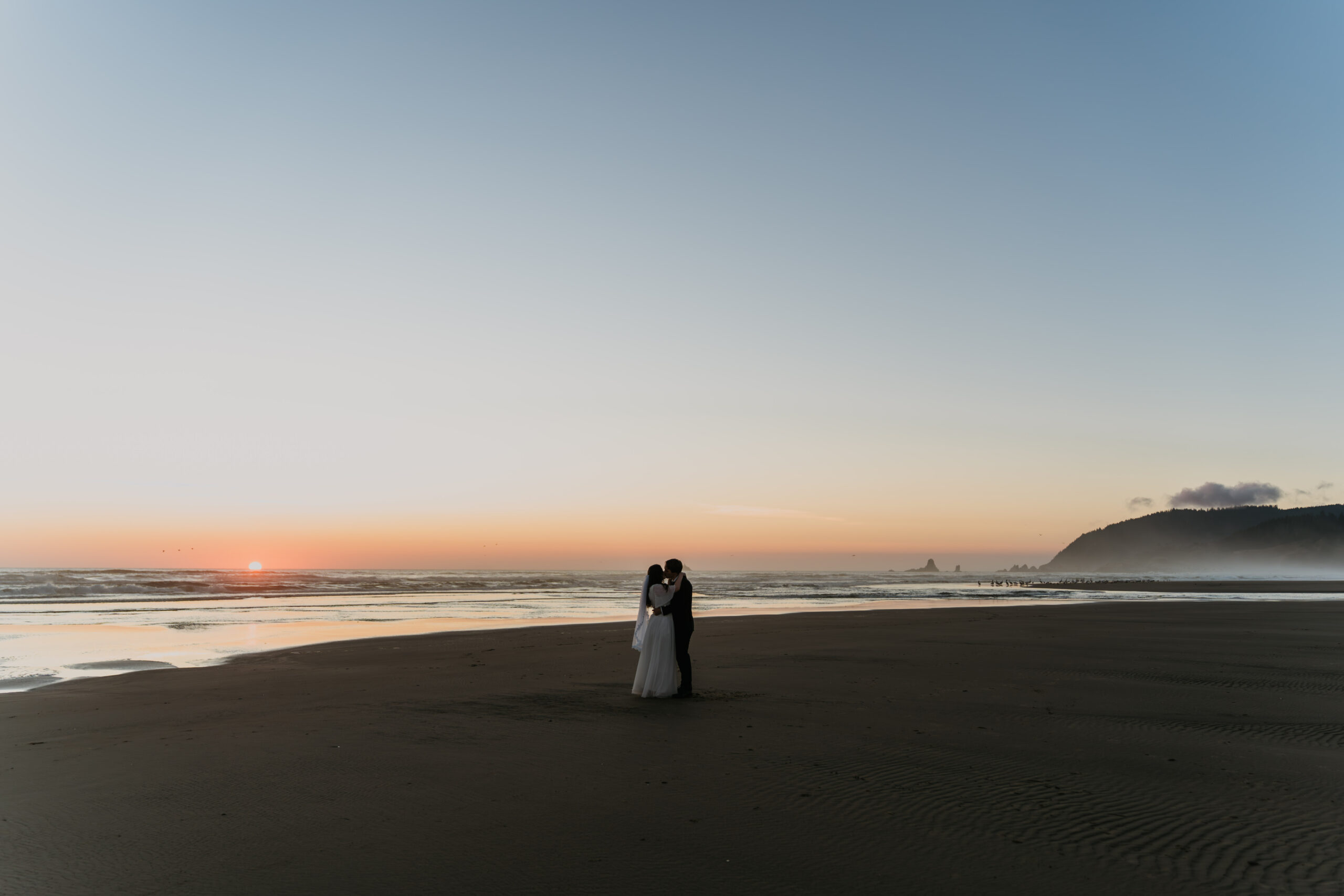 bride and groom standing together on the beach with the sunset barely showing with no one around