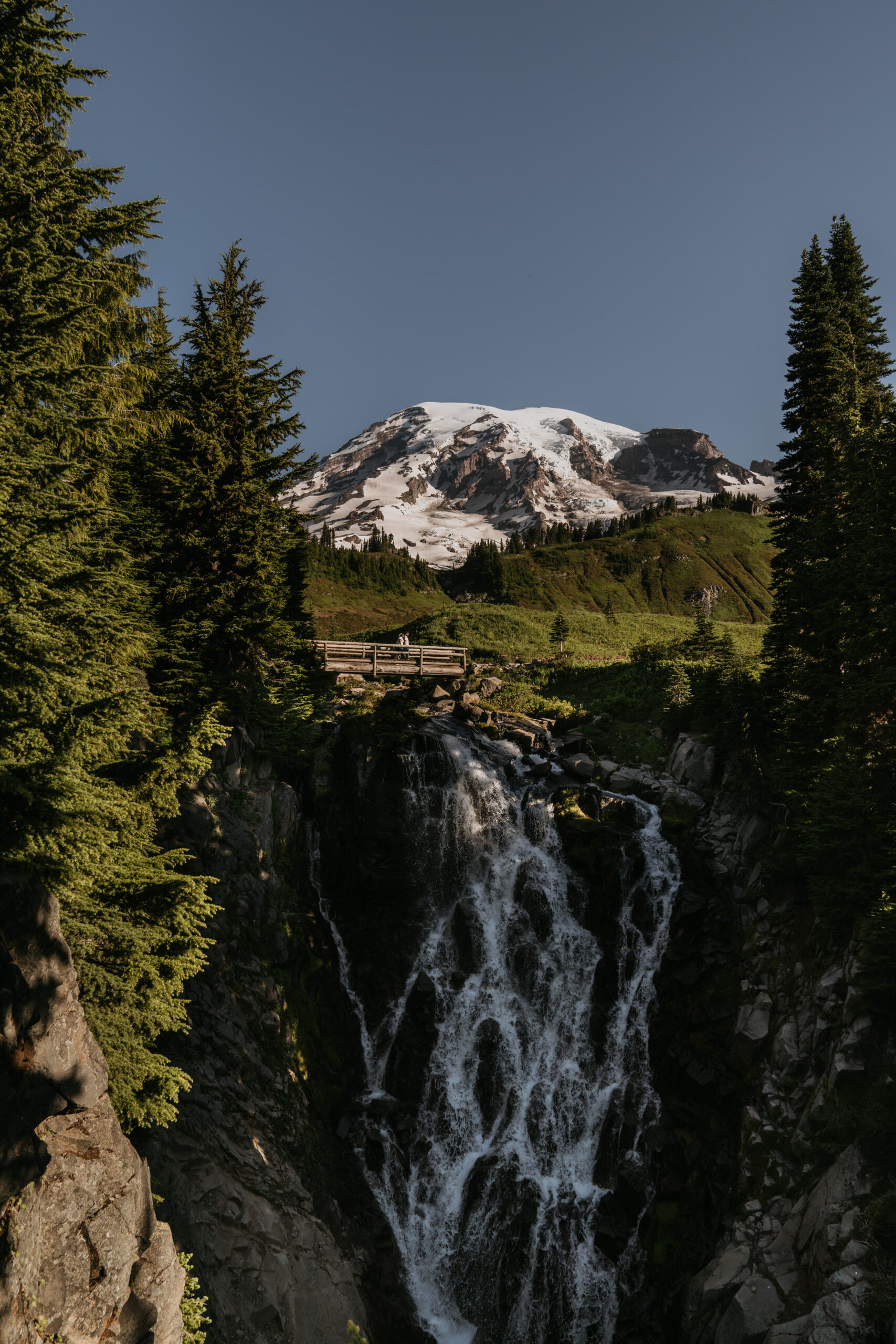 couple in far distance walking on bridge over massive waterfall with big mountain in backdrop surrounded by trees