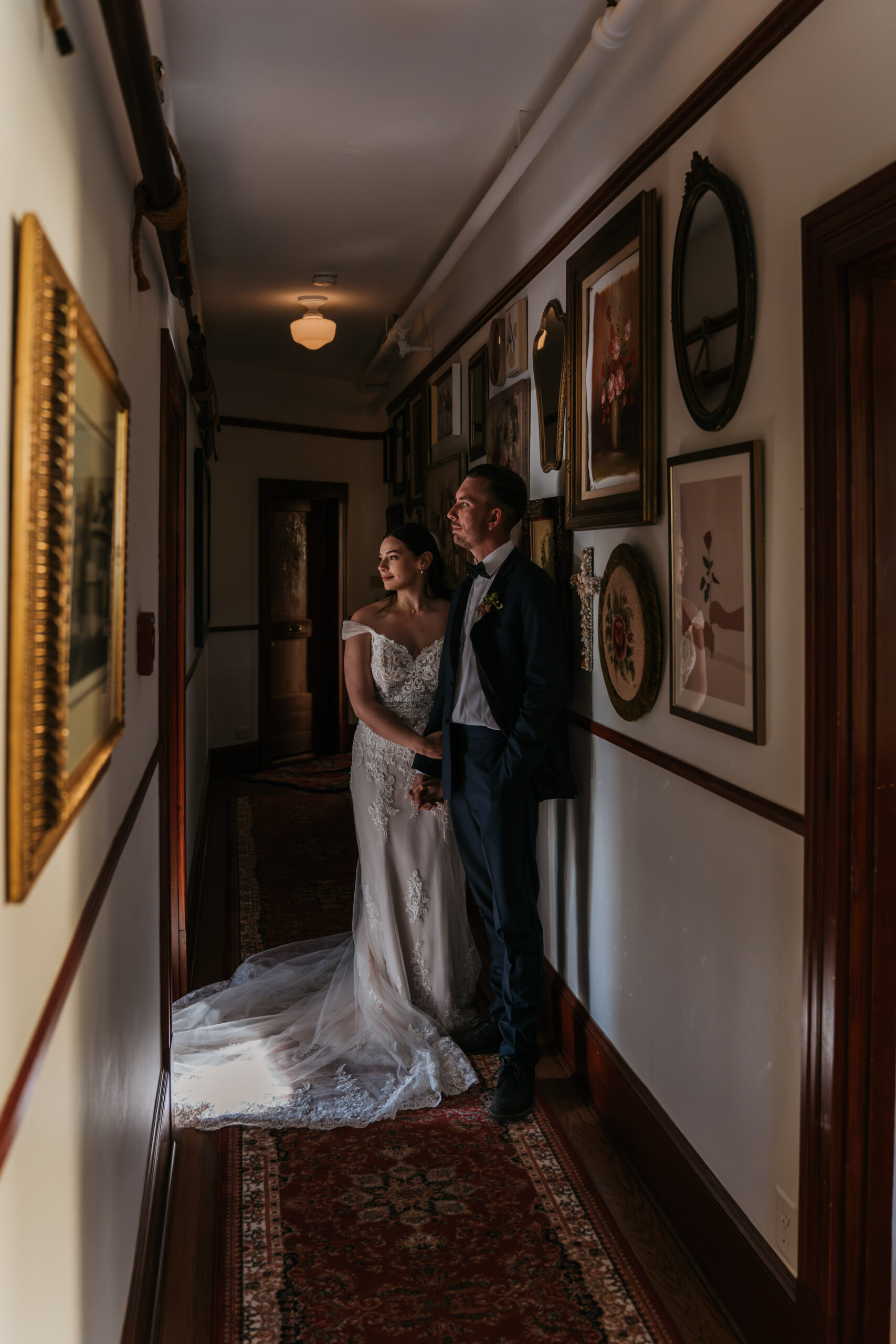 bride and groom standing in hallway looking away with wall full of antique frames and decor