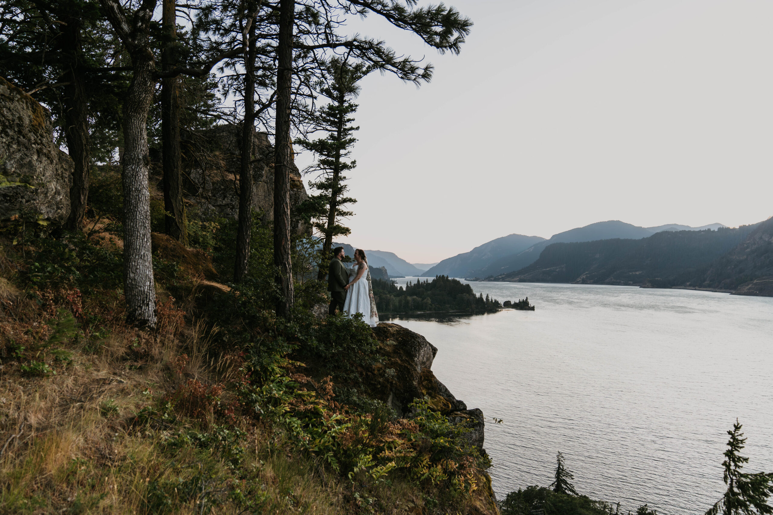 couple in distance standing on rocky cliff with massive mountain backdrop with river