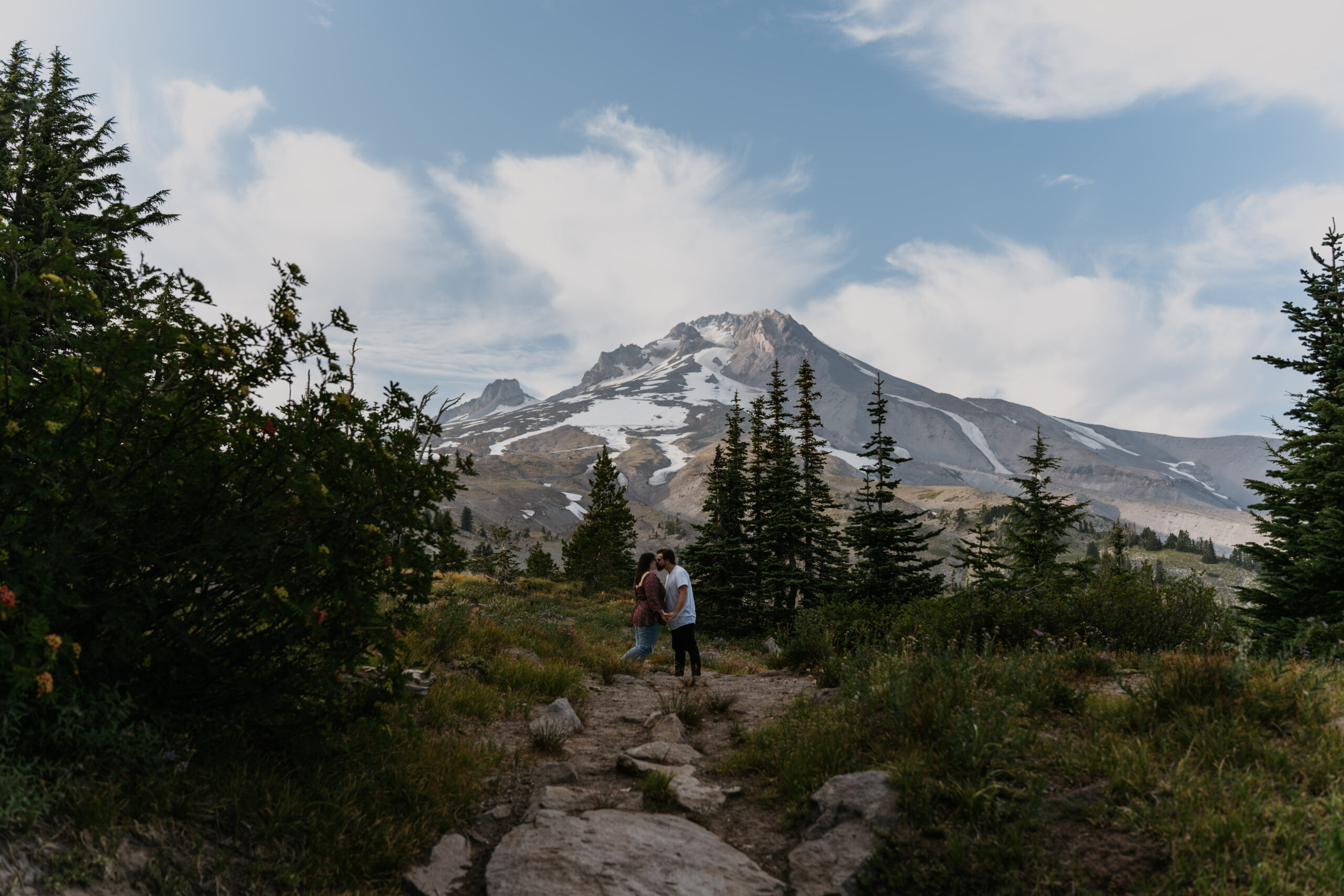 couple standing in distance in front of big mountain with forest surrounding the pathway