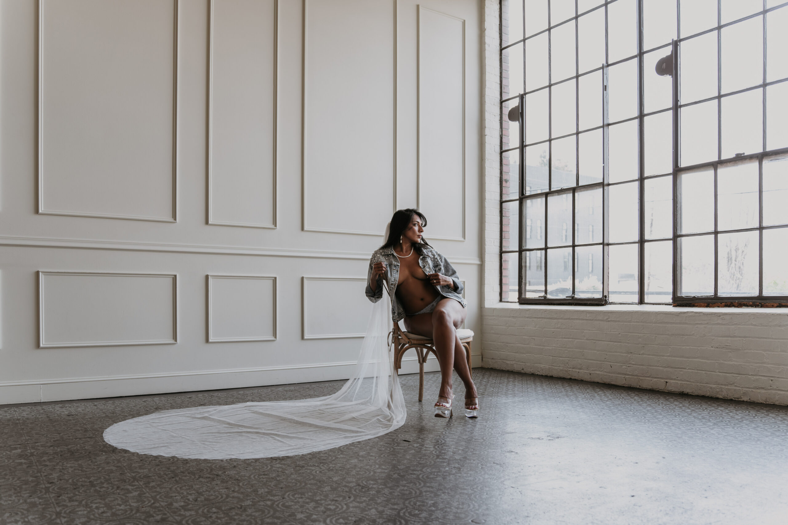 women sitting in chair in jean jacket with veil on in open room of windows for bridal boudoir session