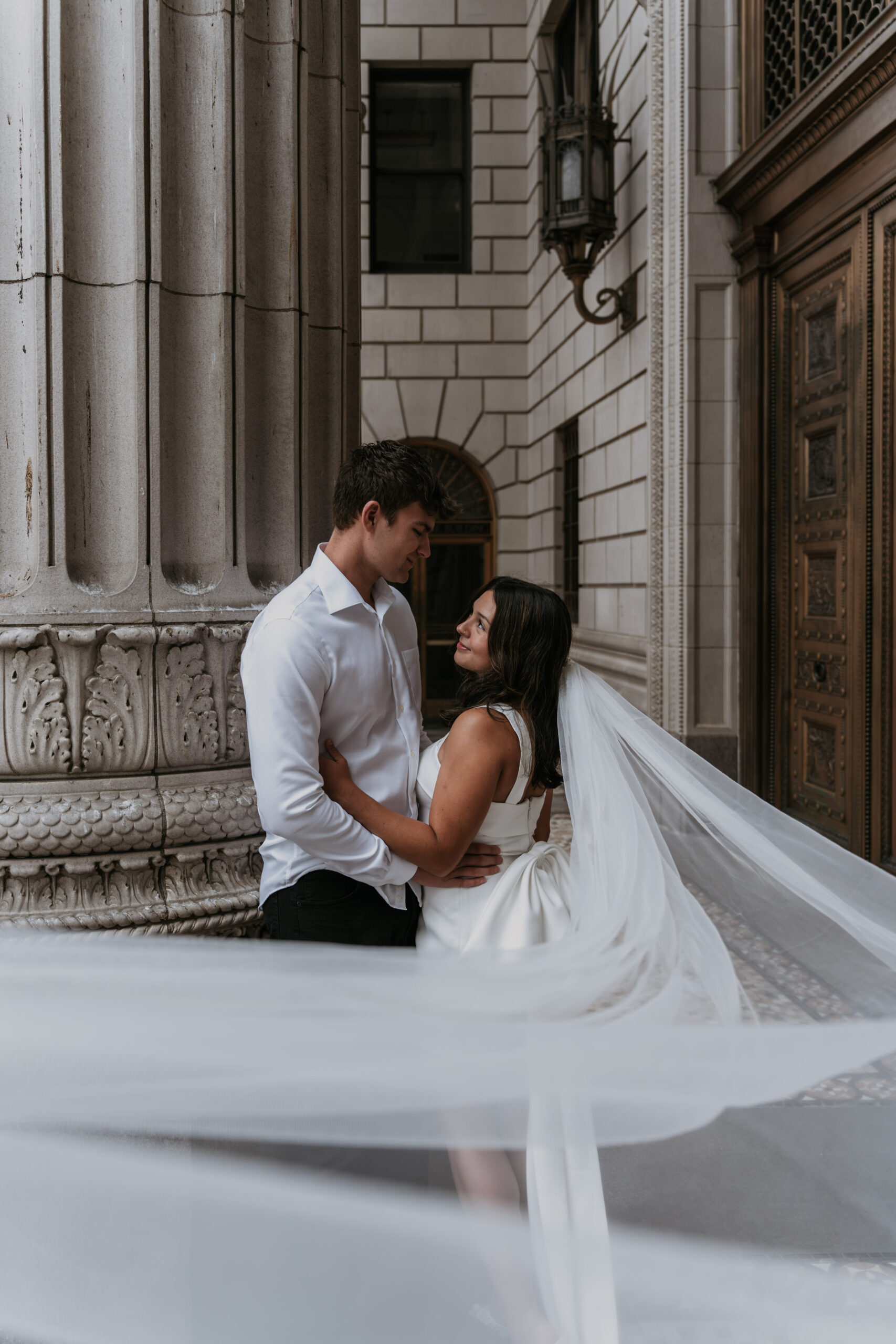 man and women holding each other in front of ancient pillars wearing bridal dress with bow and long veil