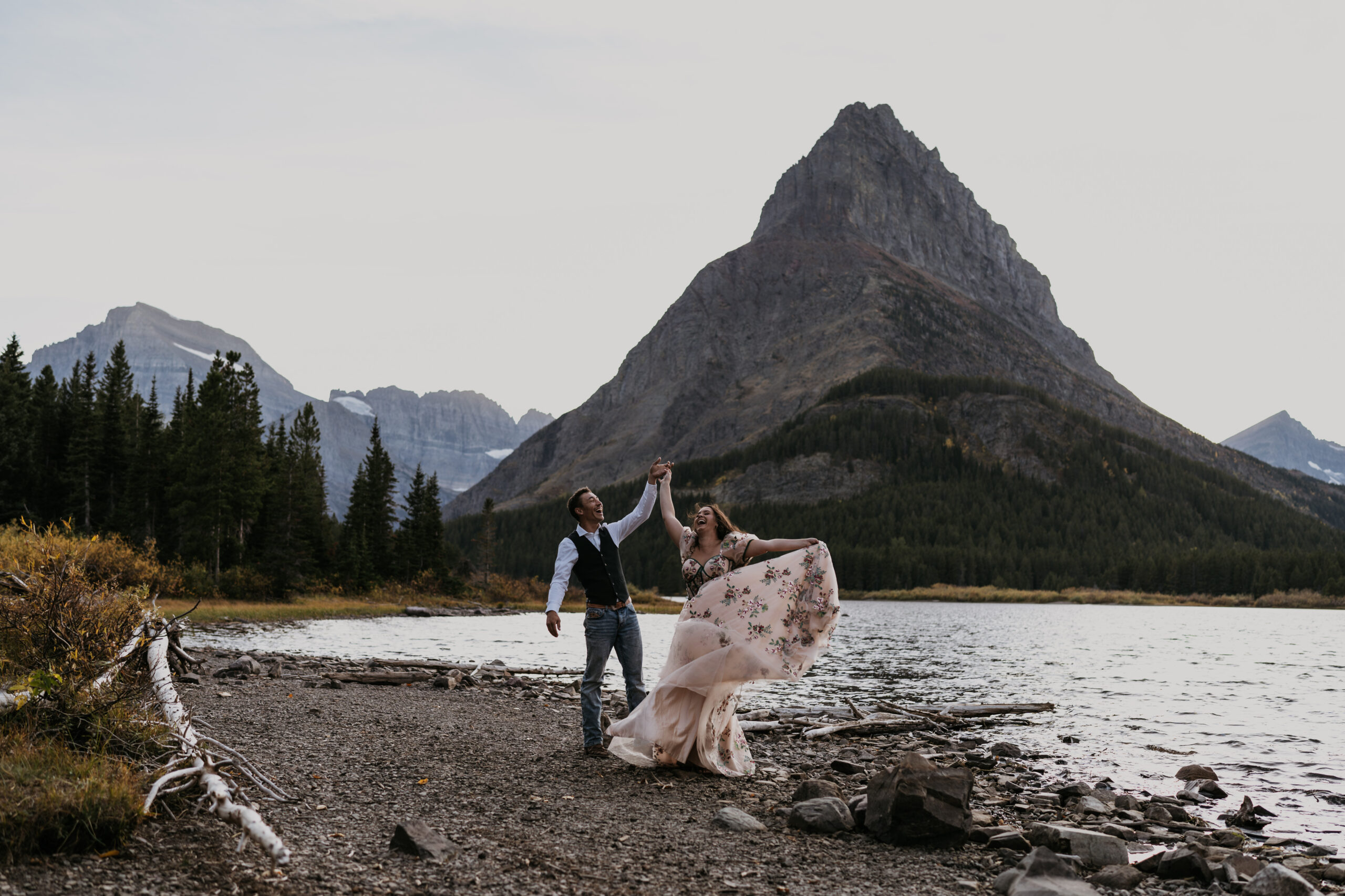 bride and groom spinning around laughing in front of big mountain by lake