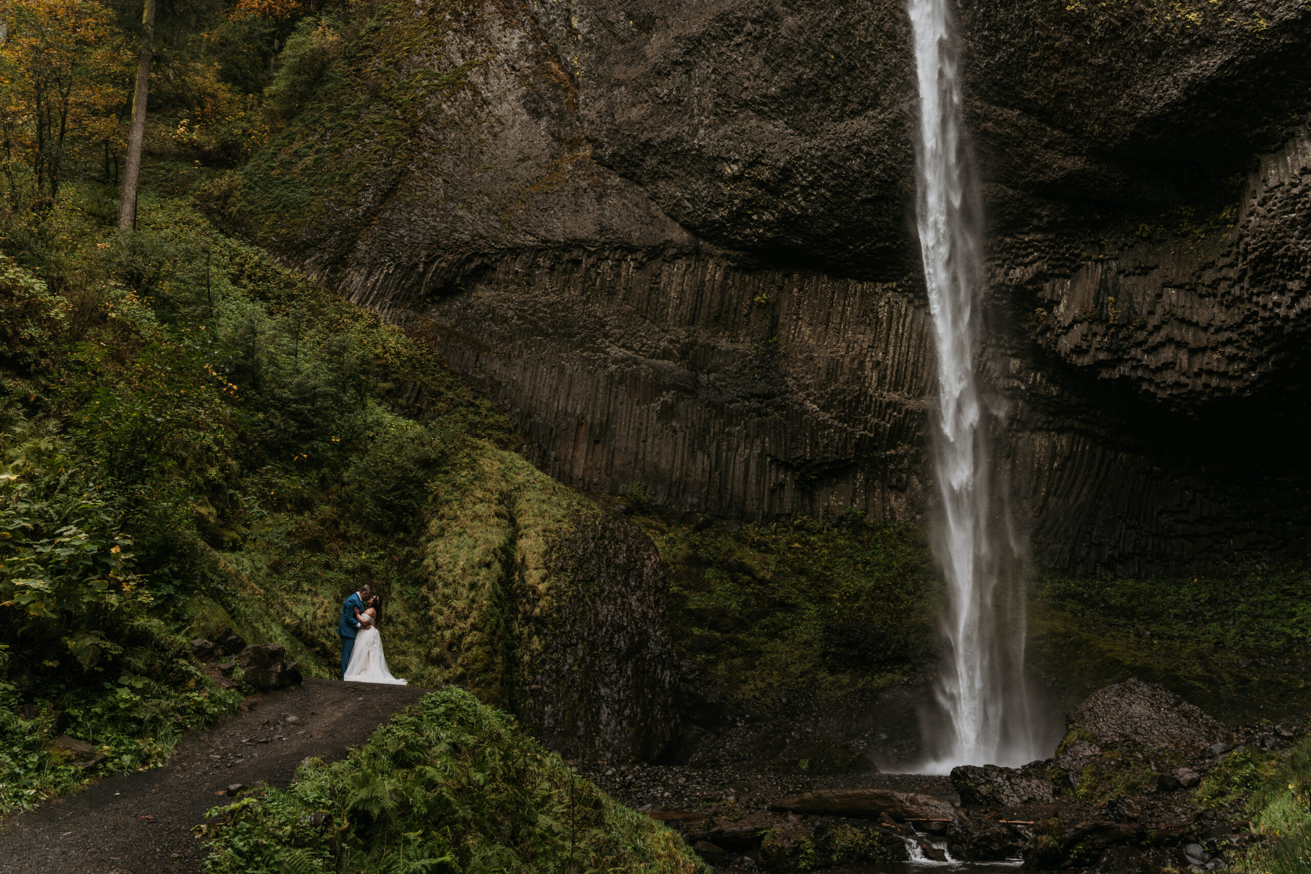 bride and groom standing in front of waterfall surrounded by lush greenery plants