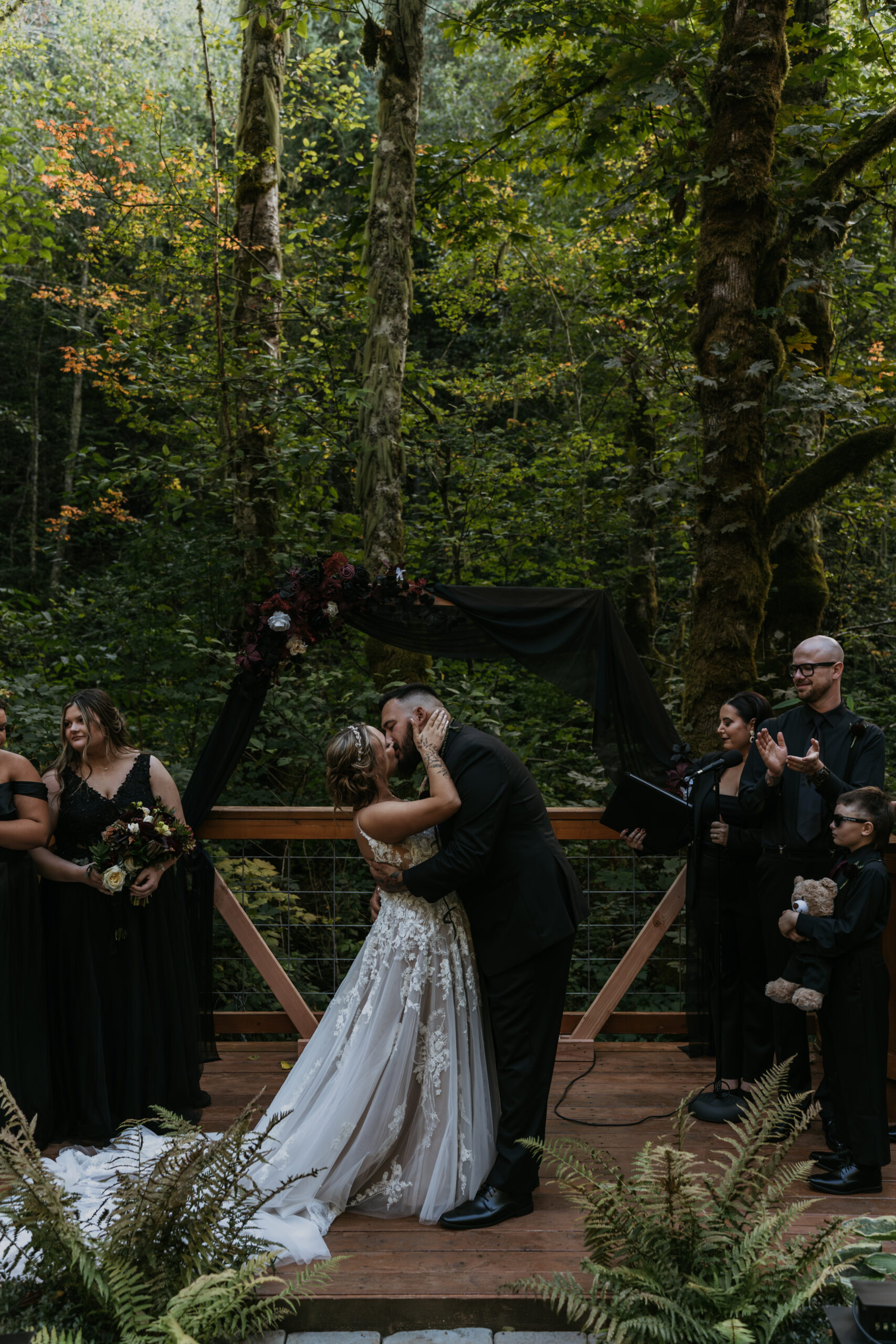 bride and groom at alter kissing with forest background with wedding party in all black