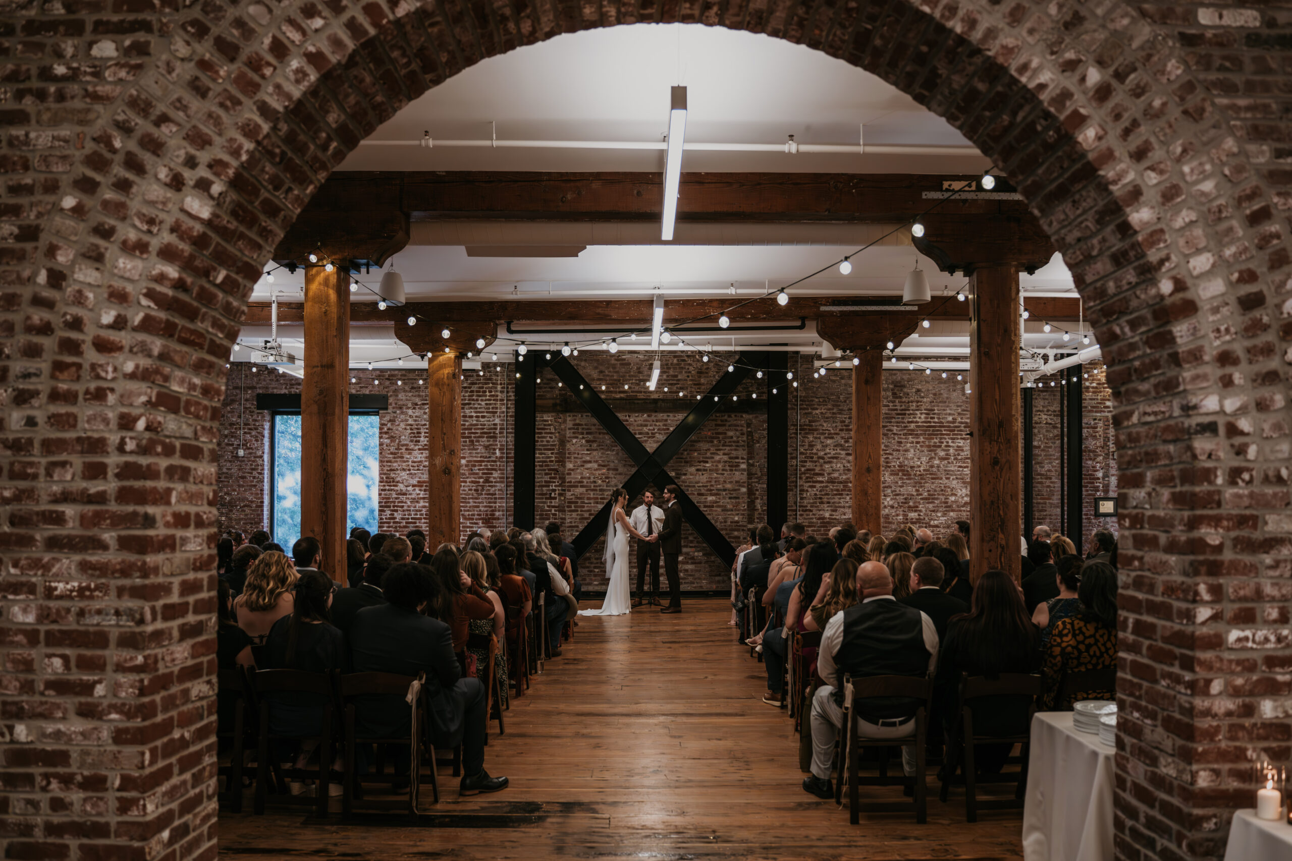 a bride and groom standing at alter in a brick building with wedding guests sitting in chair