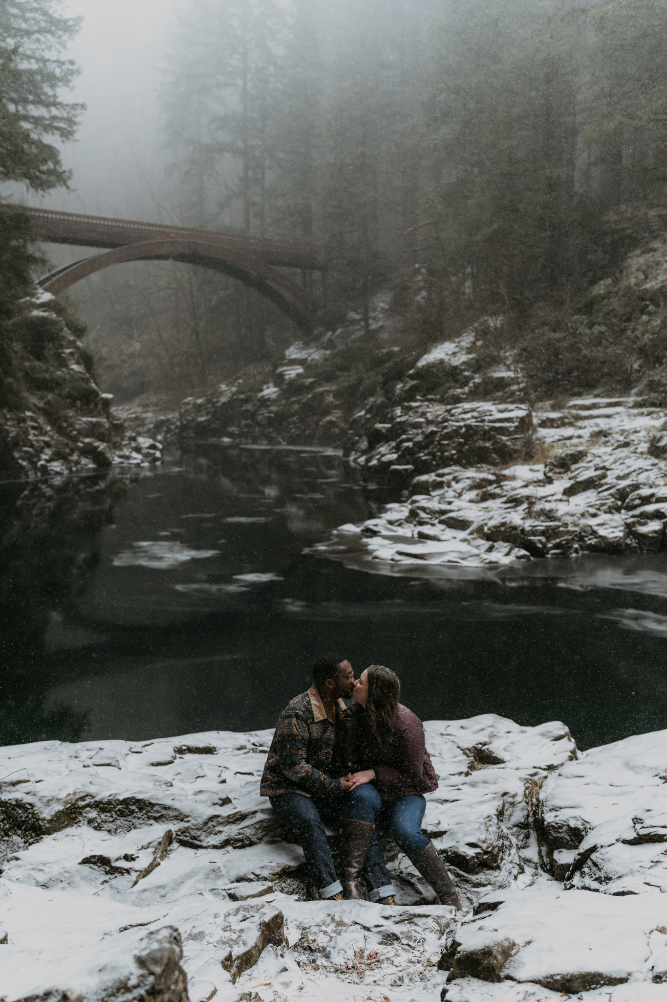 man and women sitting in snow with river and bridge and trees behind them giggling
