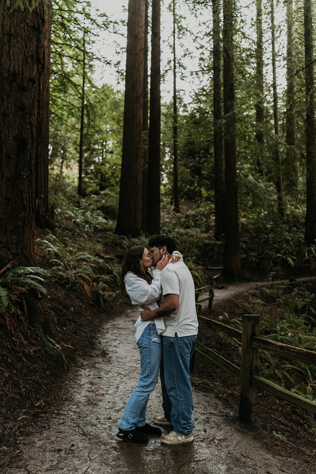 couple kissing on forest trail with tall trees surrounding them