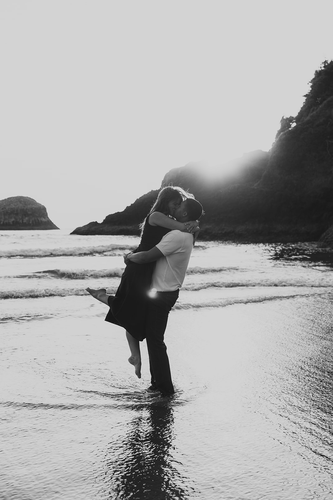 man lifting women up in ocean on beach with sunset behind rocky backdrop