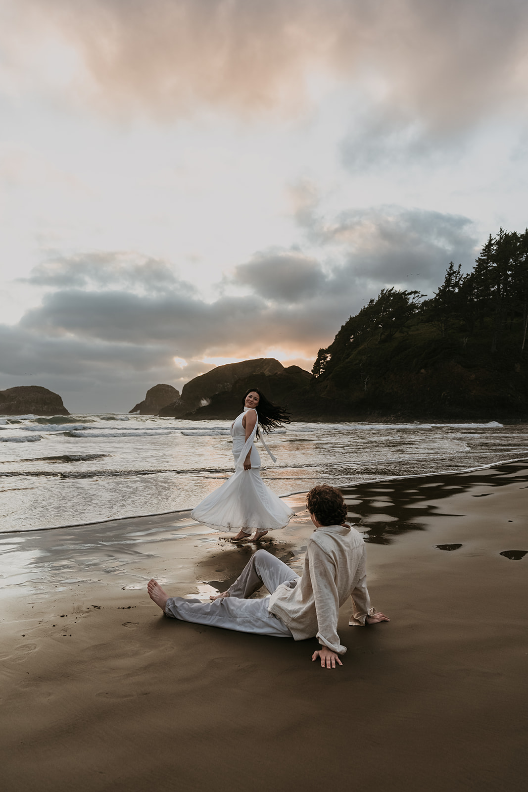 women running around twirling on beach during sunset while man sits on sand watching her
