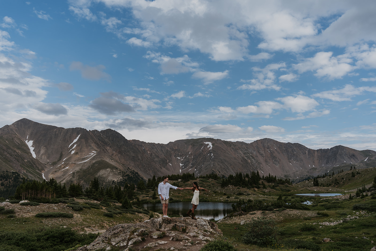 women and man holding hands on rock with big mountain lake background with blue skies and clouds