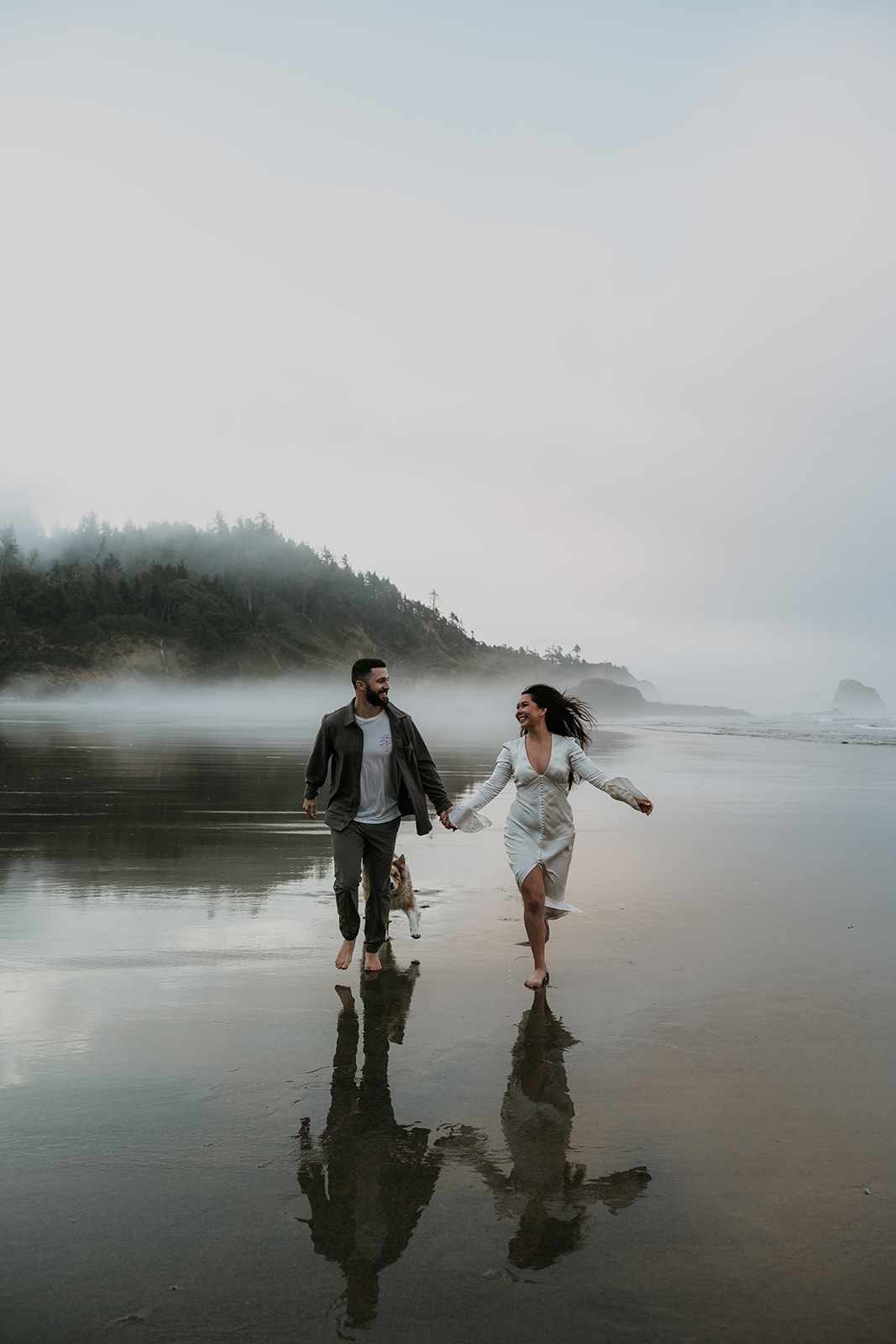 couple running on beach with fog and trees behind them, and you can see their reflection on the wet sand