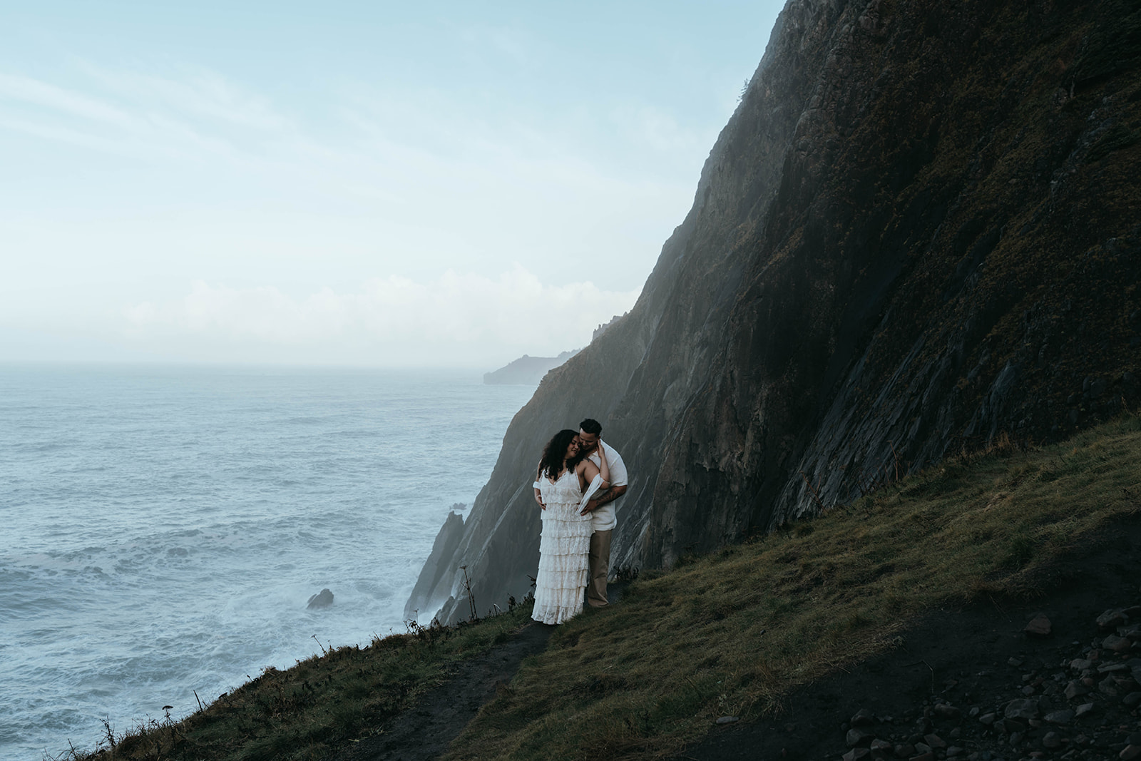 man and women holding each other in front of rocky cliff with blue skies and waves crash below