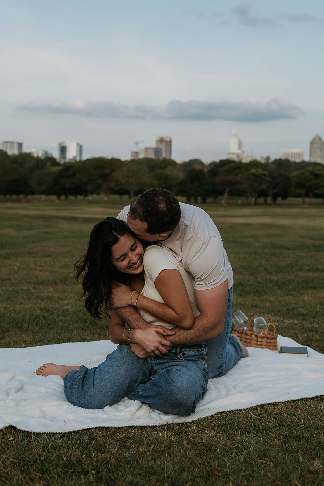 man hugging women from behind kissing her cheek and she's laughing sitting on a picnic blanket with city in background