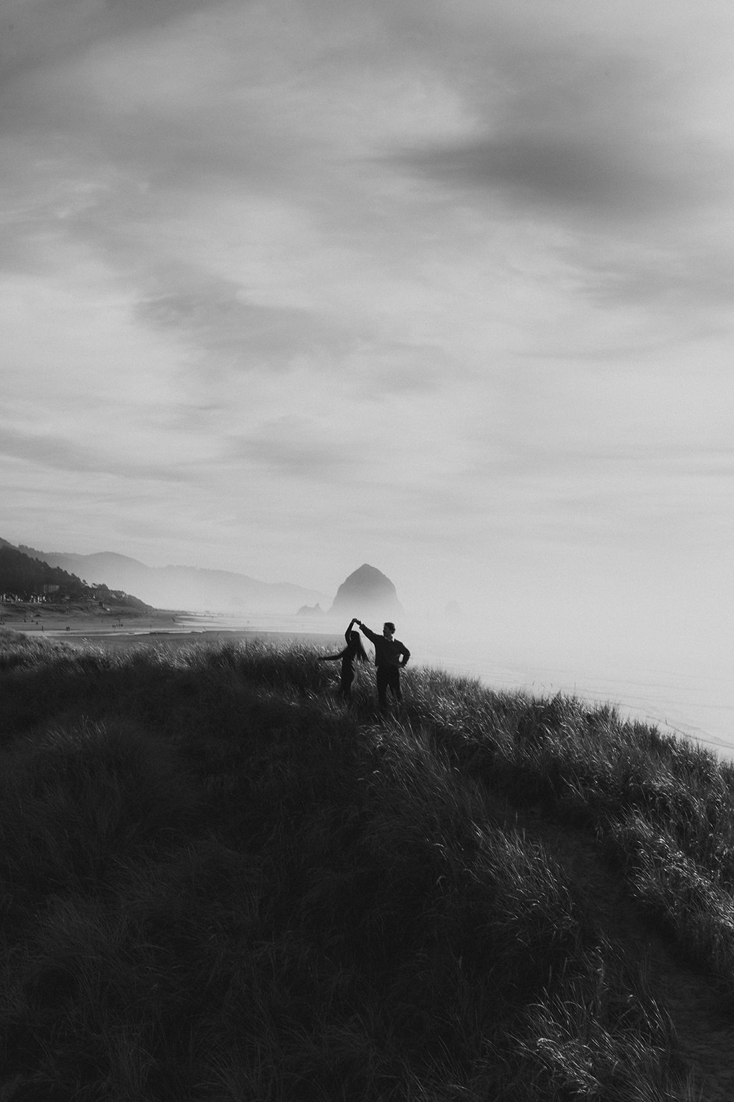 man spinning women around in beach grass on a hill with a huge rock in the background on a cloudy day