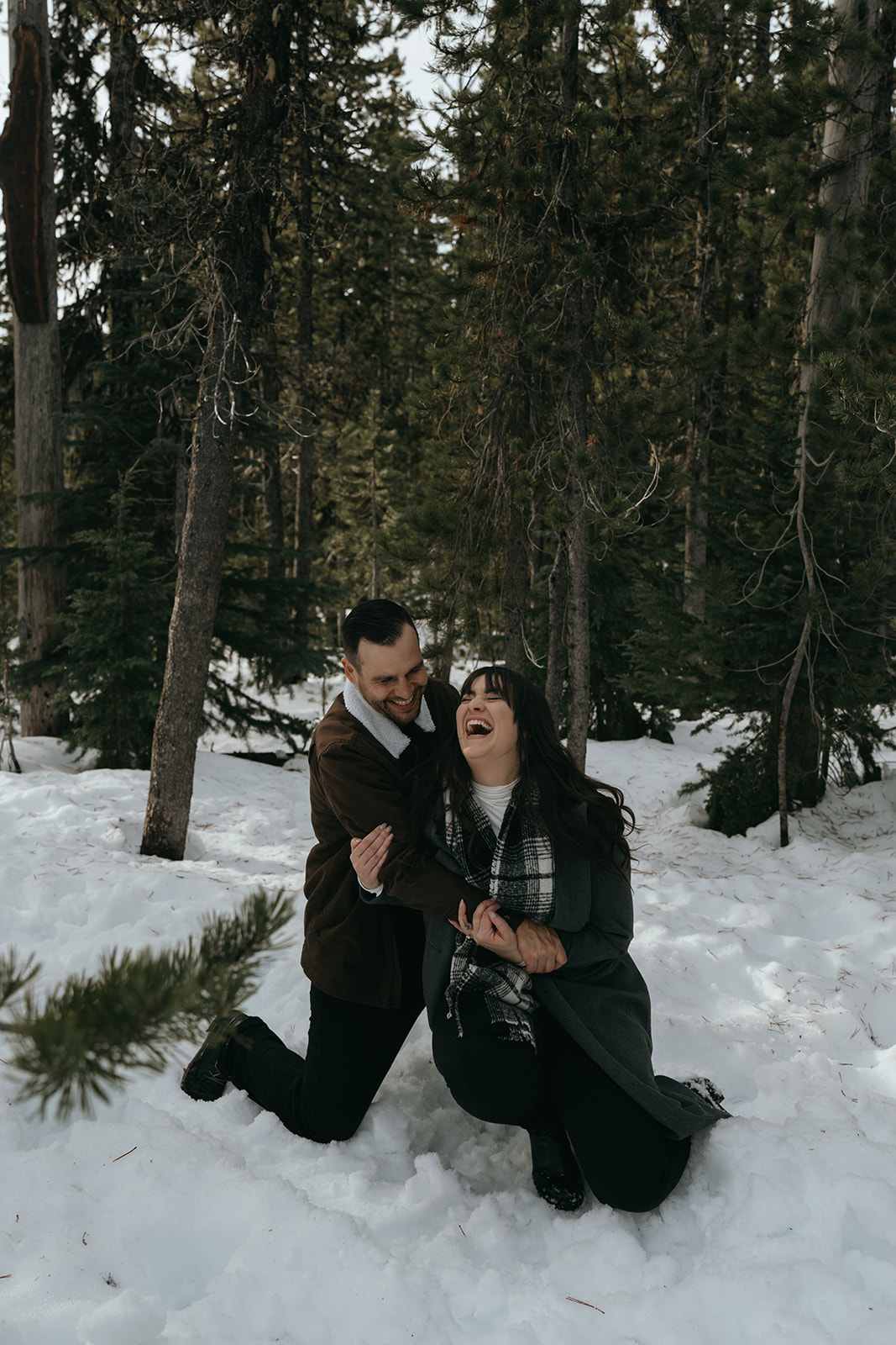 man and women laughing hard holding each other falling down in a forest with snow on the ground