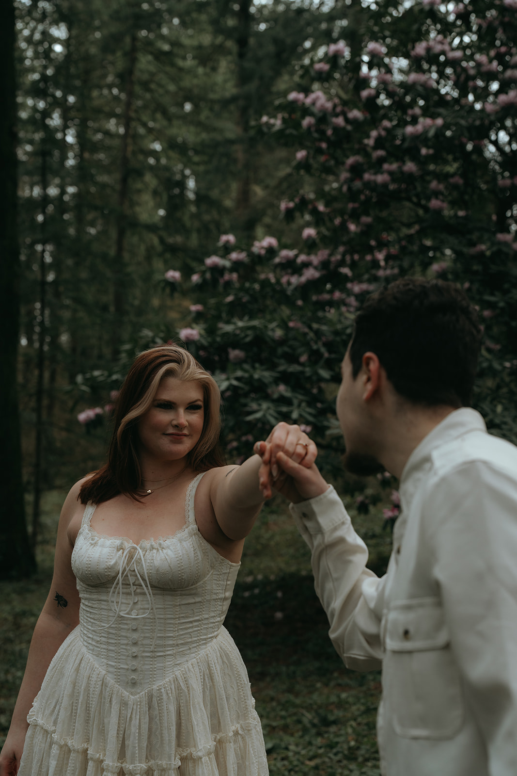 man about to kiss women's hand while women in Lacey white dress in forest with flowers behind her