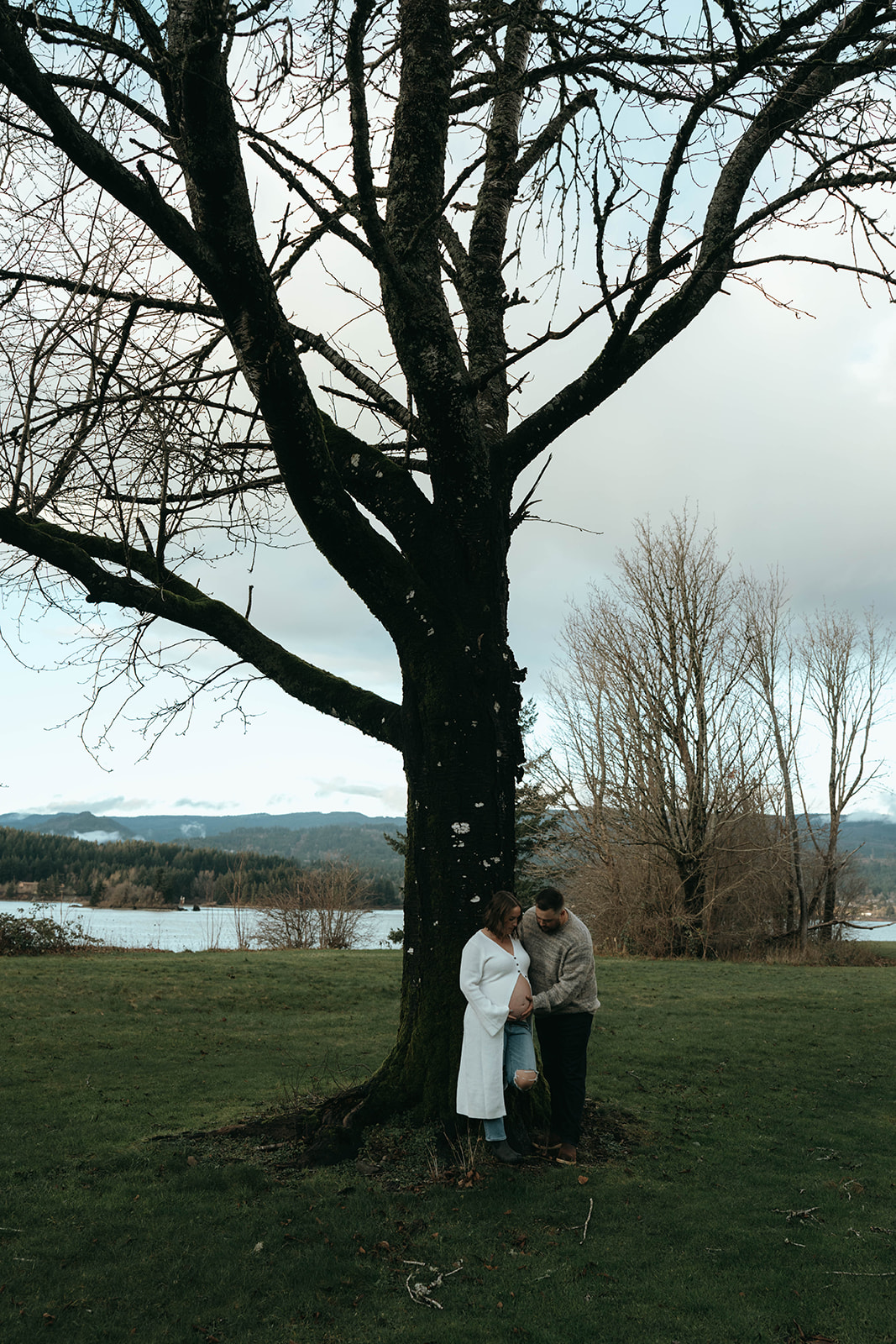 man and pregnant women holding each other next to tree in grassy field with river and mountains in background