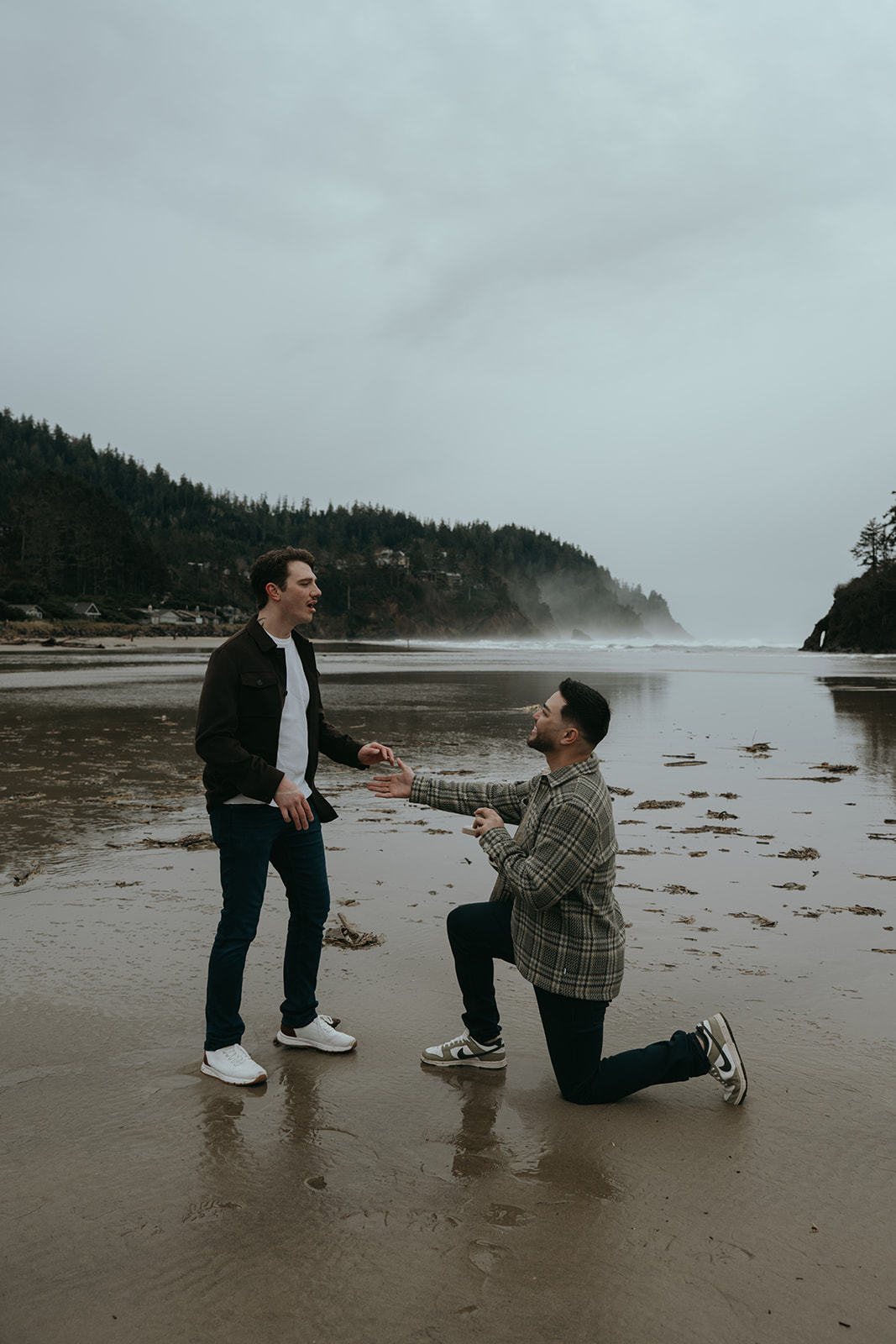man proposing to man on beach with cloudy skies and forest in background on the wet sad, man in shock