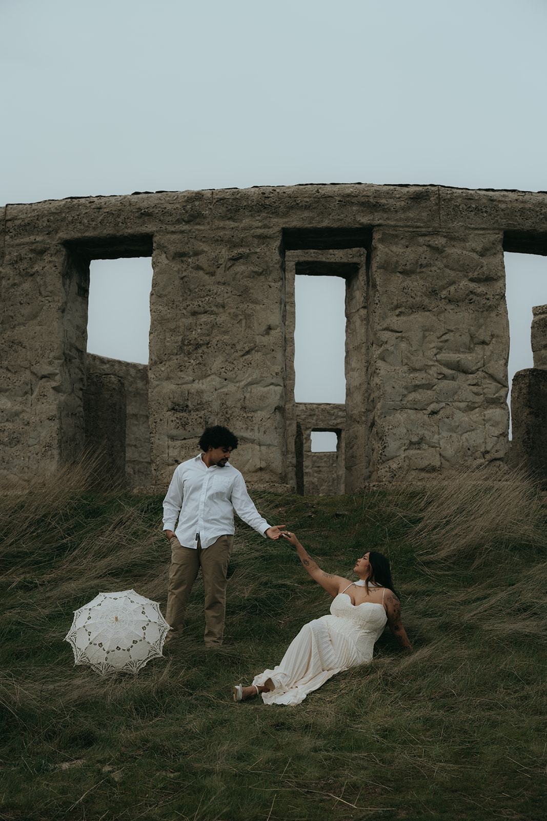 bride sitting on grass reaching out at groom while groom is standing reaching out at bride, in front of a Stonehenge on a cloudy day