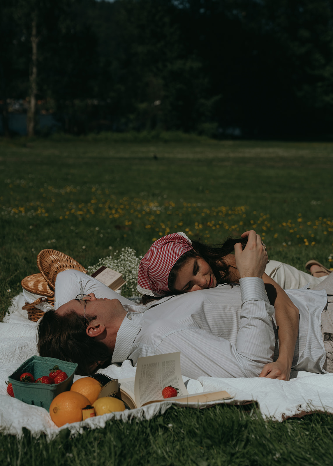 women laying on man's chest while he plays with her hair, on a picnic blanket with fruits all around them