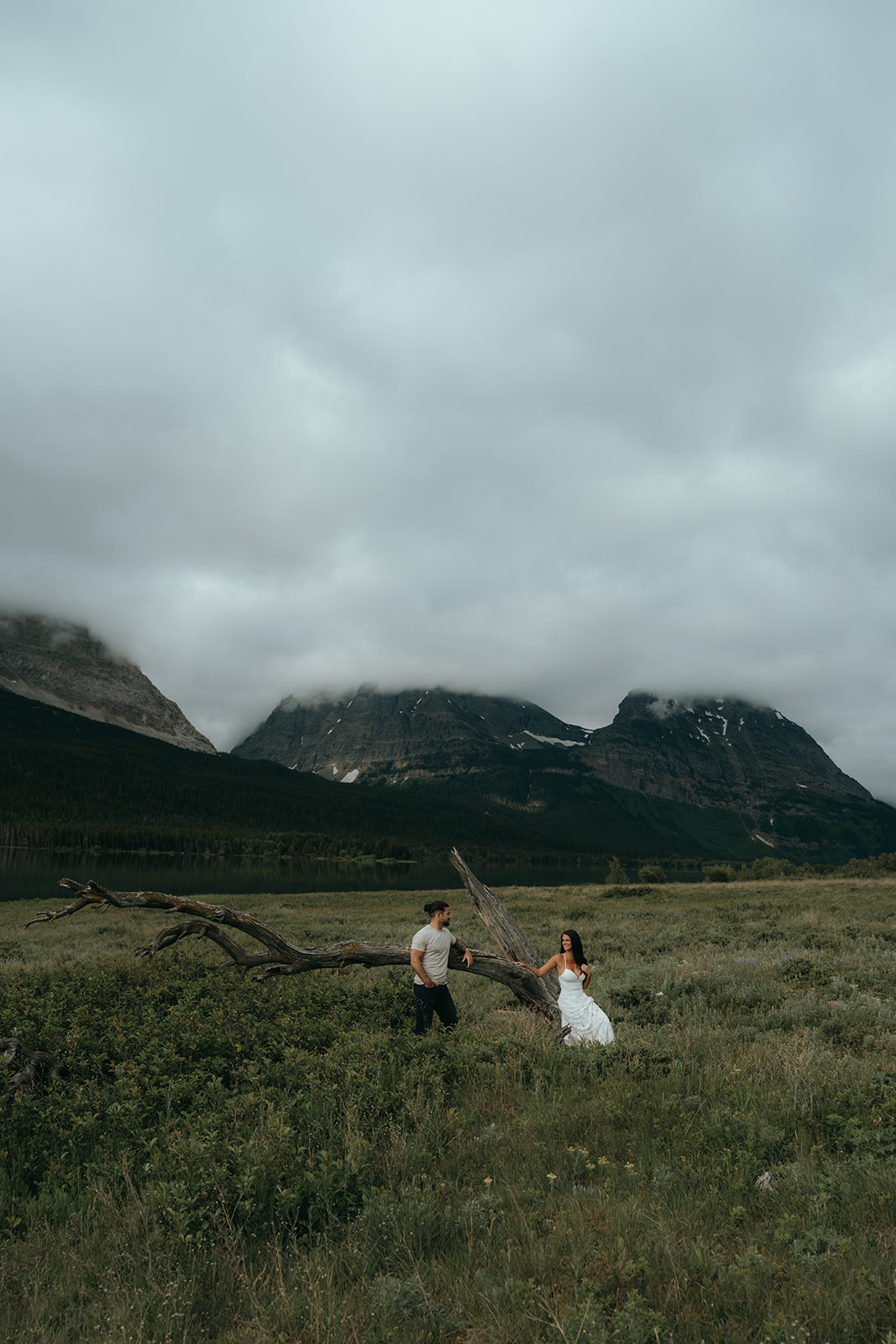 man and women sitting on old tree in grassy field with huge mountains in the background on a cloudy day
