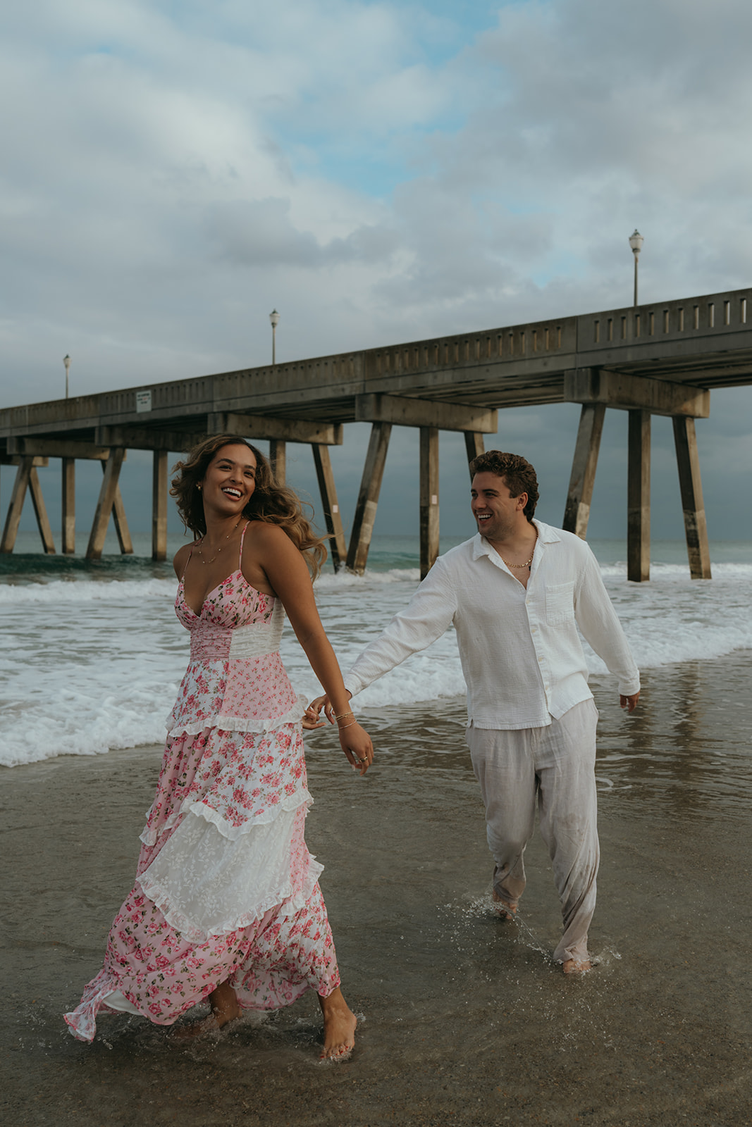 man and women laughing on beach running around in ocean water with bridge behind them on a blue cloudy day