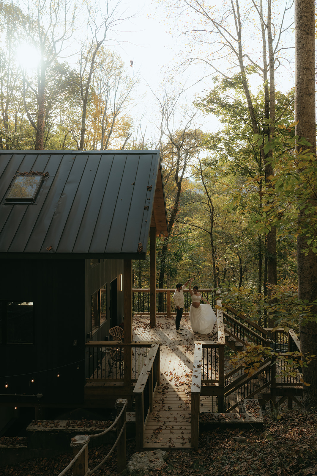 man spinning women around on a deck attached to a black cabin in the woods during fall with the sun shining through