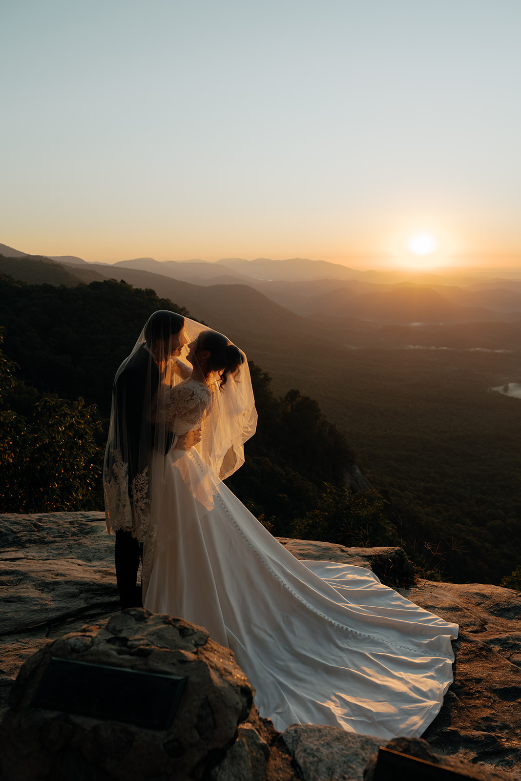 bride and groom standing on rock viewpoint with mountains as background and golden sun peaking through over the hills